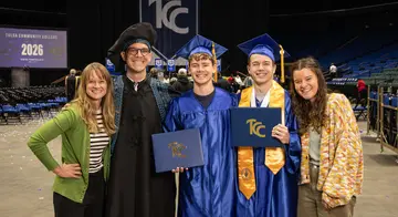Ashley, Professor Da Corte, Hudson, Everett, and Breese pose for a photo and smile. Hudson and Everett are wearing blue graduation regalia and hold their degrees.
