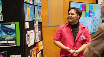 Digital Media student Keith Ponce speaks with spectators about his work. He is looking at his portfolio which is displayed on a wall.