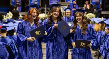 Three women dressed in blue graduation cap and gowns hold up TCC degrees