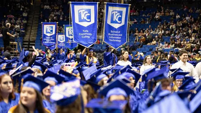 Two students in blue graduation regalia each hold a TCC gonfalon in a crowd of graduating students
