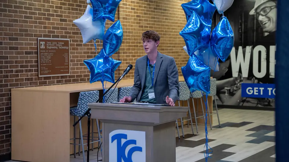 Student Alex Jones speaks behind a podium flanked by blue and white balloons inside the Engineering Technology Building.