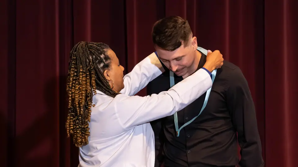 Faculty Dr. Angela Elmore drapes a ribbon with a Nursing pin around a student's neck. The student is a man wearing a black button-up shirt.