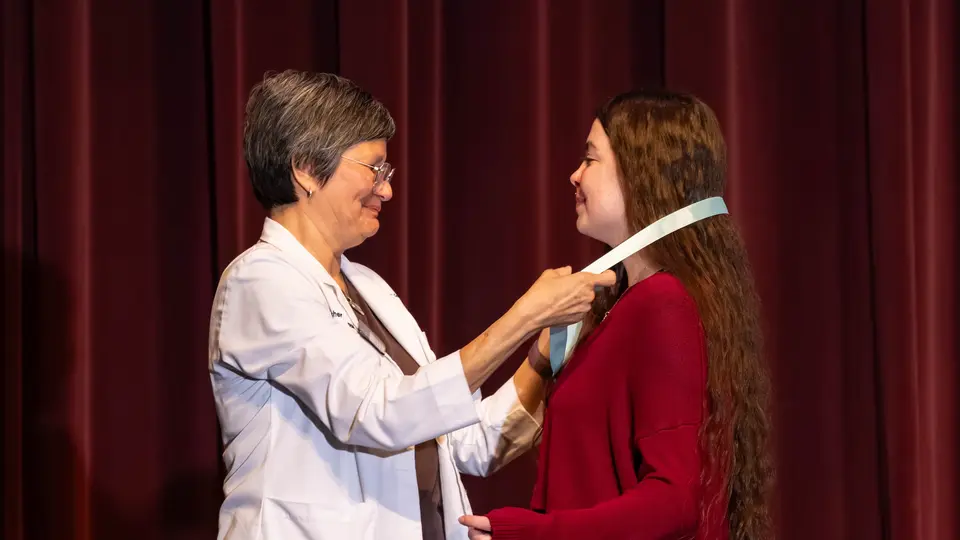 Faculty Angela Archer drapes a ribbon with a Nursing pin around a student's neck. The student is a woman wearing a red dress.