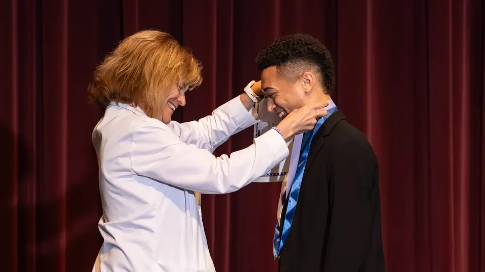 Faculty Susan Goldsmith drapes a ribbon with a Nursing pin around a student's neck. The student is a man wearing a black button-up shirt. Both people are smiling.