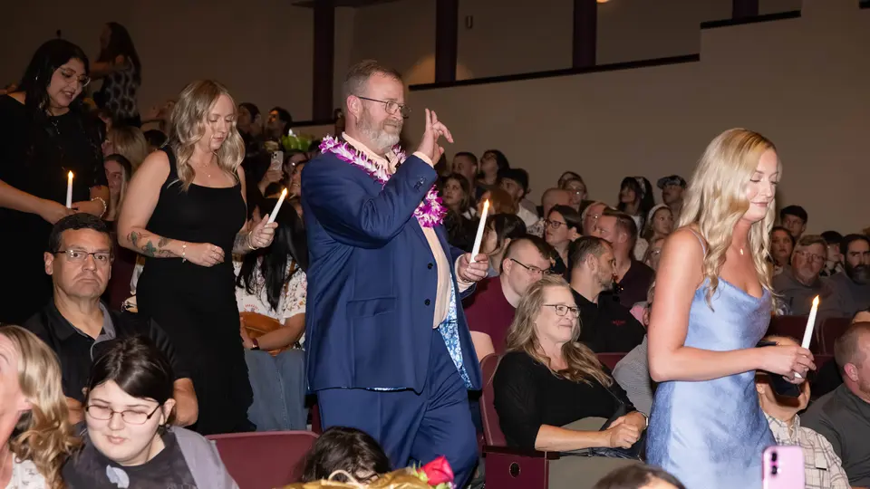Students each holding one white candle walk down stairs in the VanTrease PACE to be seated for the ceremony. A man in a blue suit with a pink Hawaiian lay is giving the peace sign to someone in the crowd.