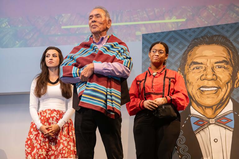 Ivy Collins, Wes Studi and Kianle Frazier stand on stage after a blanket presentation