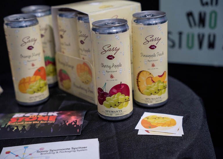 MaryAnn Ferrell's design on a mock can of spumante spritzer. There are six can visible, and all are a light yellow color. They have designs of various fruits on them.