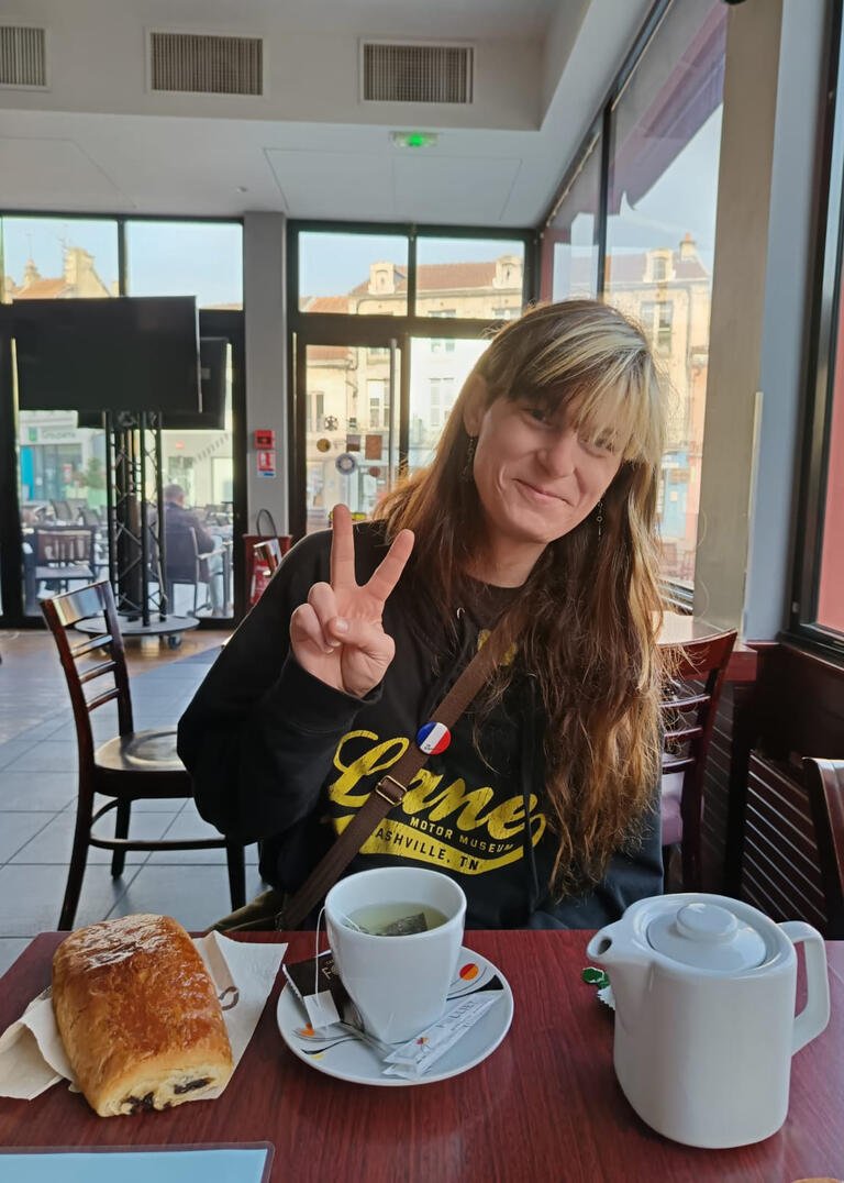 Natasha gives the peace sign at a cafe in France. She is sitting at a table with a cup of coffee and a pastry sitting on the table in front of her.